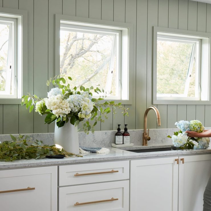 Bright kitchen with open awning windows, fresh flowers, and a woman arranging blooms at the sink